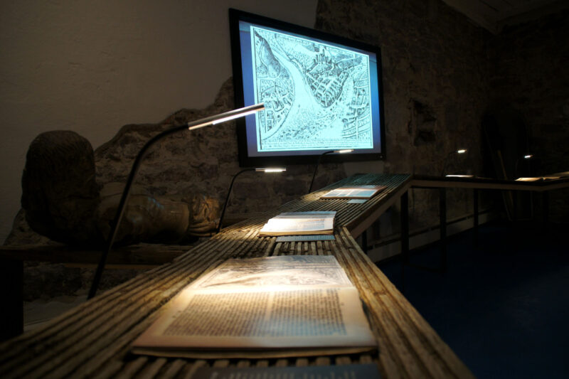 Ausstellungsansicht im Grafschaftsmuseum Wertheim. Auf einem Bildschirm siht man eine historische Karte, auf einem Tisch liegen historische Schriften. Das Thema ist Hochwasser in Wertheim am Main.
